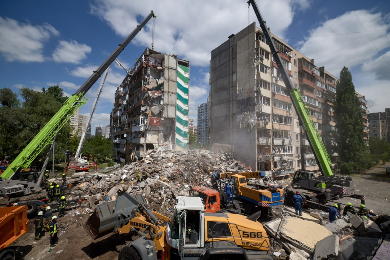 Рятувальники та важка техніка розбирають завали зруйнованої дев'ятиповерхівки. Два великі зелені підйомні крани та екскаватор Liebherr працюють посеред уламків бетону між двома пошкодженими житловими будинками під сонячним небом із хмарами.