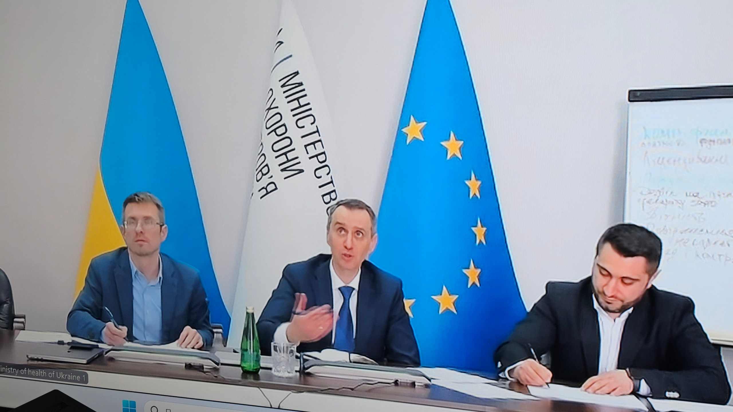 Офіційна онлайн-нарада за участю трьох представників у ділових костюмах, які сидять за столом перед камерою. На задньому плані встановлені державний прапор України, прапор Європейського Союзу та білий стяг із написом «Міністерство охорони здоров'я».