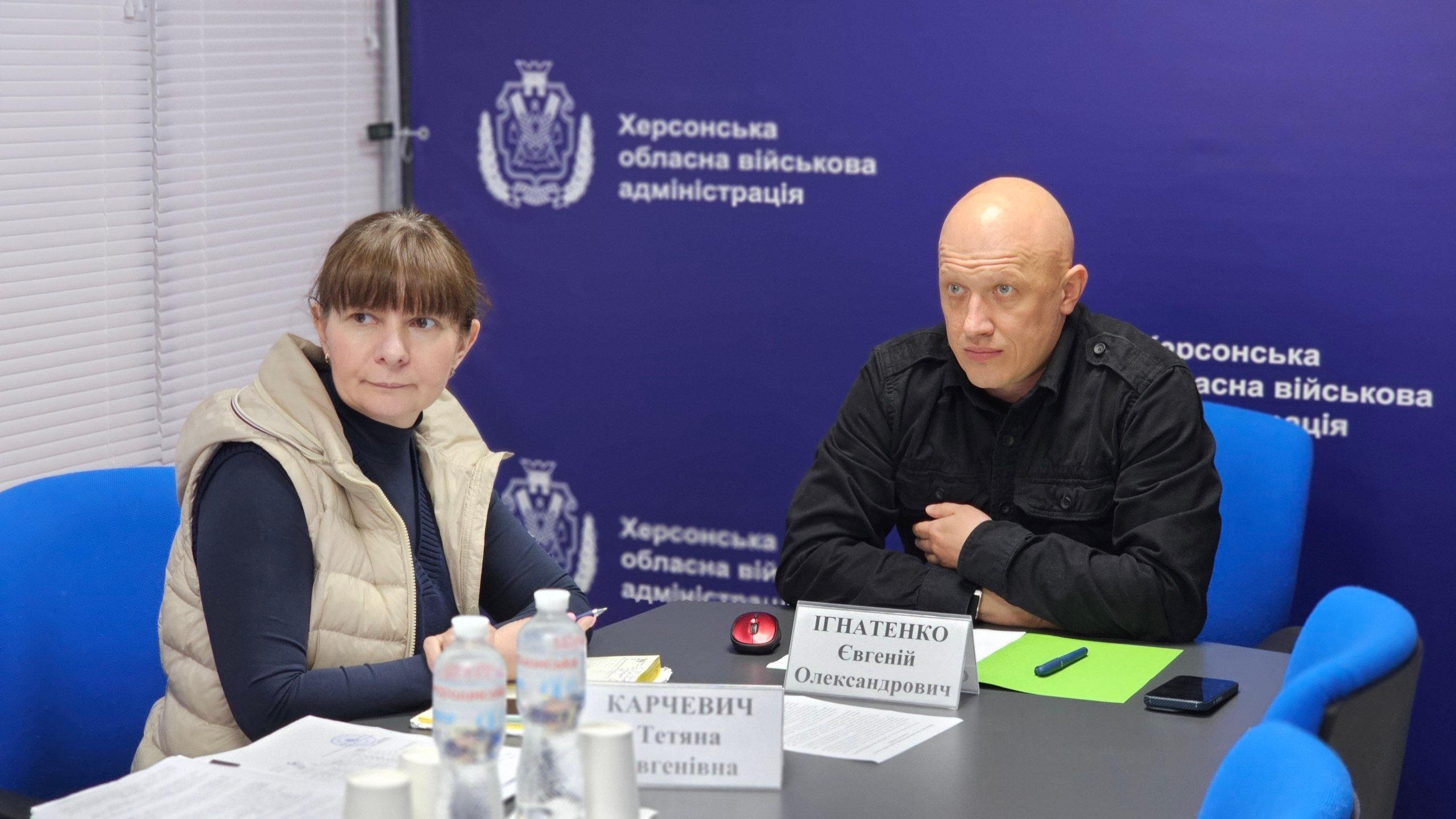 Робоча зустріч двох представників за столом в офіційному приміщенні. Перед ними розташовані іменні таблички, папери, блокноти та пляшки з водою, а на синьому банері позаду видно герби та текстові написи.