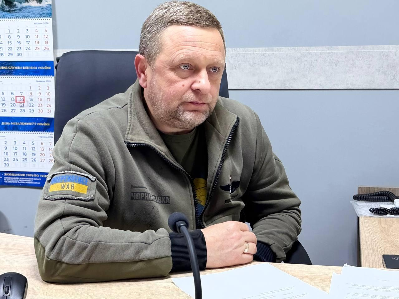 Чоловік у військовій кофті кольору хакі з нашивкою у вигляді прапора України та написом «WAR» сидить за робочим столом. На столі перед ним стоїть мікрофон та лежать документи, а ліворуч на стіні висить настінний календар.