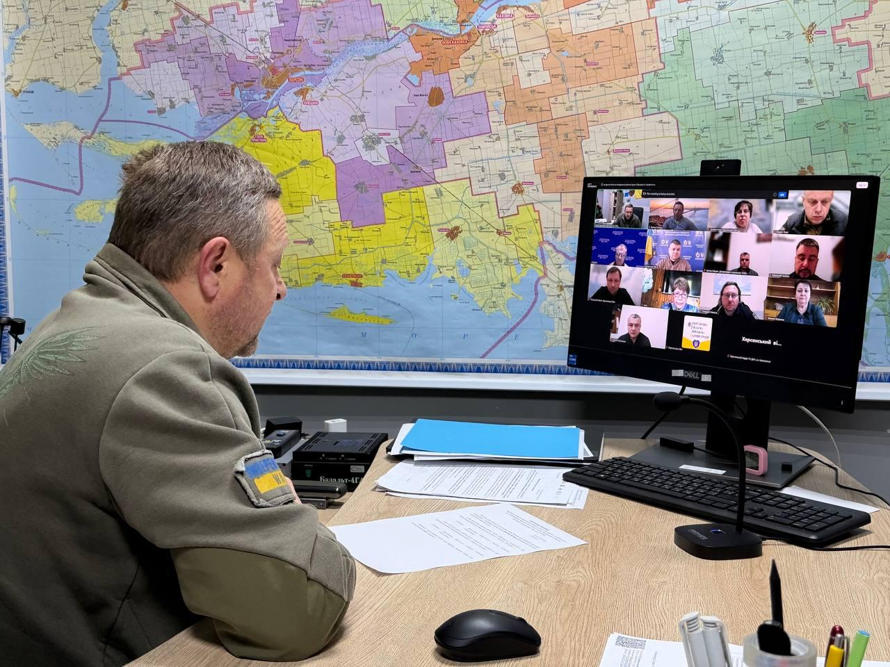 Чоловік у військовій кофті кольору хакі сидить за робочим столом і бере участь у відеоконференції на великому моніторі. Перед ним лежать документи та клавіатура, а на стіні позаду комп'ютера висить масштабна кольорова карта місцевості.