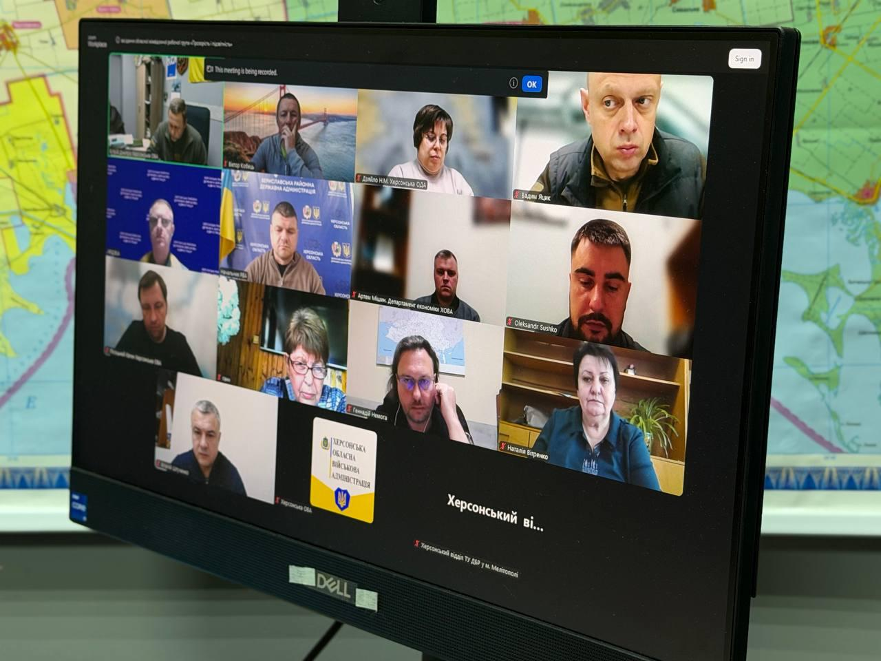 Монітор комп'ютера Dell, на якому відображено вікно онлайн-наради у форматі відеоконференції з багатьма учасниками. В одному з вікон на чорному тлі видно офіційну емблему та герб, а позаду монітора розміщена велика географічна карта.