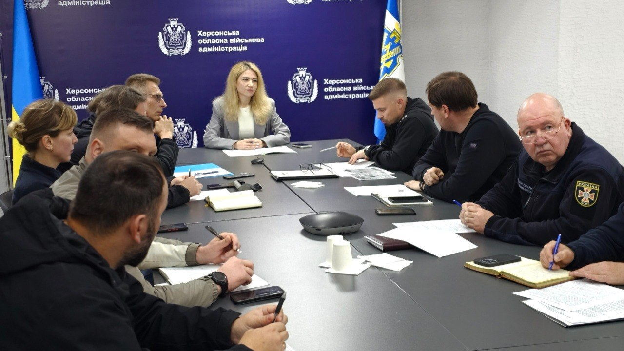 Група людей проводить робочу нараду за довгим сірим столом у офіційному приміщенні. Учасники сидять по обидва боки столу з документами та блокнотами, а на синьому банері позаду видно державні прапори, офіційні емблеми, герби та текстові написи.