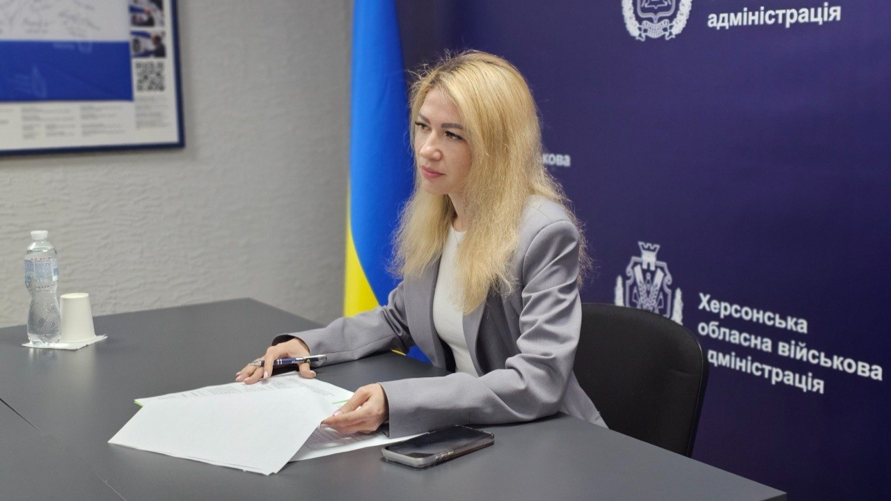 Жінка у сірому піджаку сидить за робочим столом, тримаючи ручку та вивчаючи папери. Поруч лежить смартфон, стоїть пляшка води та стаканчик, а на синьому банері позаду видно офіційні емблеми, герби та текстові написи.