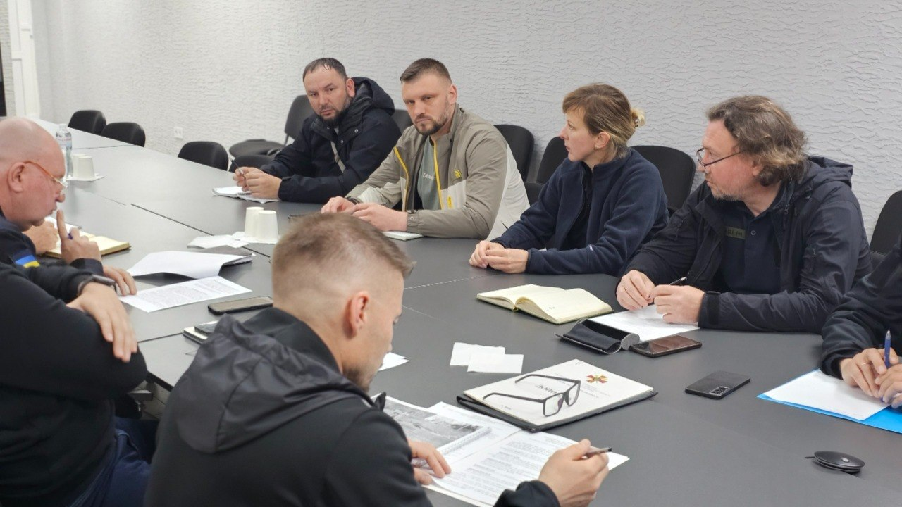 Робоча нарада учасників за великим сірим столом у кабінеті зі світлими текстурними стінами. Люди сидять навпроти один одного, вивчають документи, роблять записи в блокнотах і обговорюють робочі питання.