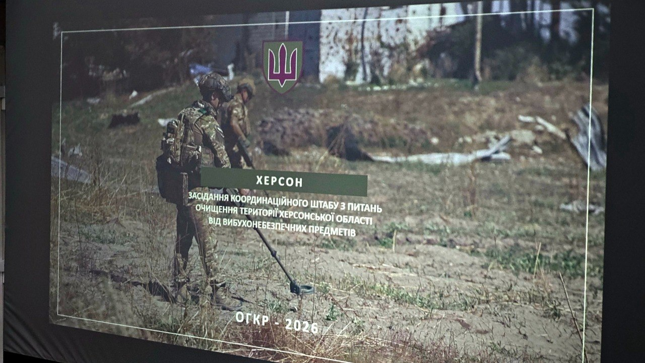 Презентаційний слайд на великому екрані з фотографією саперів у військовій формі та захисному спорядженні, які обстежують загородну місцевість металошукачами. По центру слайда розміщено малий герб України (Тризуб) та текстовий блок про засідання координаційного штабу з питань очищення території Херсонської області.