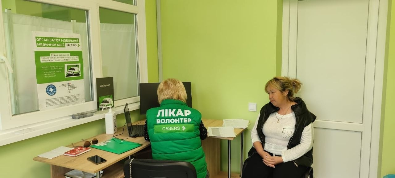 Жінка сидить на стільці біля робочого столу та спілкується з медичним працівником, який сидить спиною до кадру в зеленому жилеті з написом «ЛІКАР ВОЛОНТЕР CASERS». На столі розміщені ноутбук, документи, папки та антисептик. На скляній перегородці ліворуч висить плакат з інформацією про мобільні медичні місії.