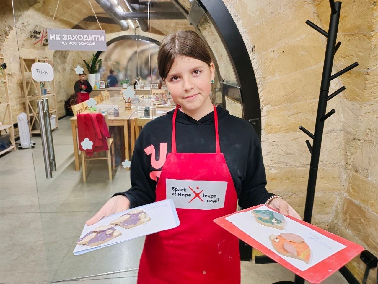Дівчинка в чорному худі та червоному фартуху з написом «Spark of Hope / Іскра надії» тримає в руках два аркуші з готовими розфарбованими виробами з глини. Вона стоїть на тлі світлої творчої майстерні з арочною стелею, де видно великий дерев'яний стіл.