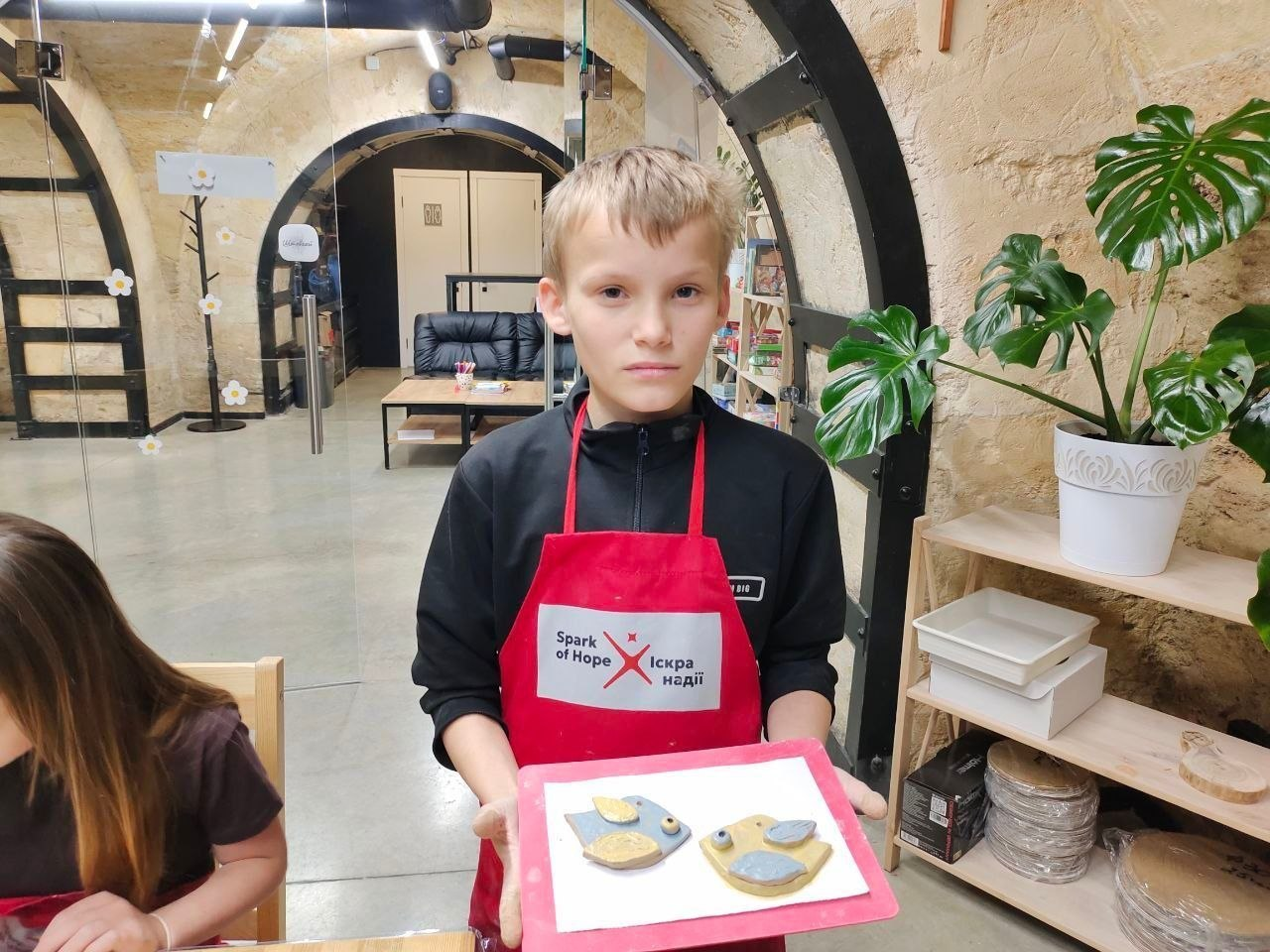 Хлопчик у чорній кофті та червоному фартуху з написом «Spark of Hope / Іскра надії» тримає в руках підставку з двома розфарбованими глиняними виробами у формі пташок. На задньому плані видно інтер'єр майстерні з арочними скляними перегородками та кімнатну рослину праворуч.