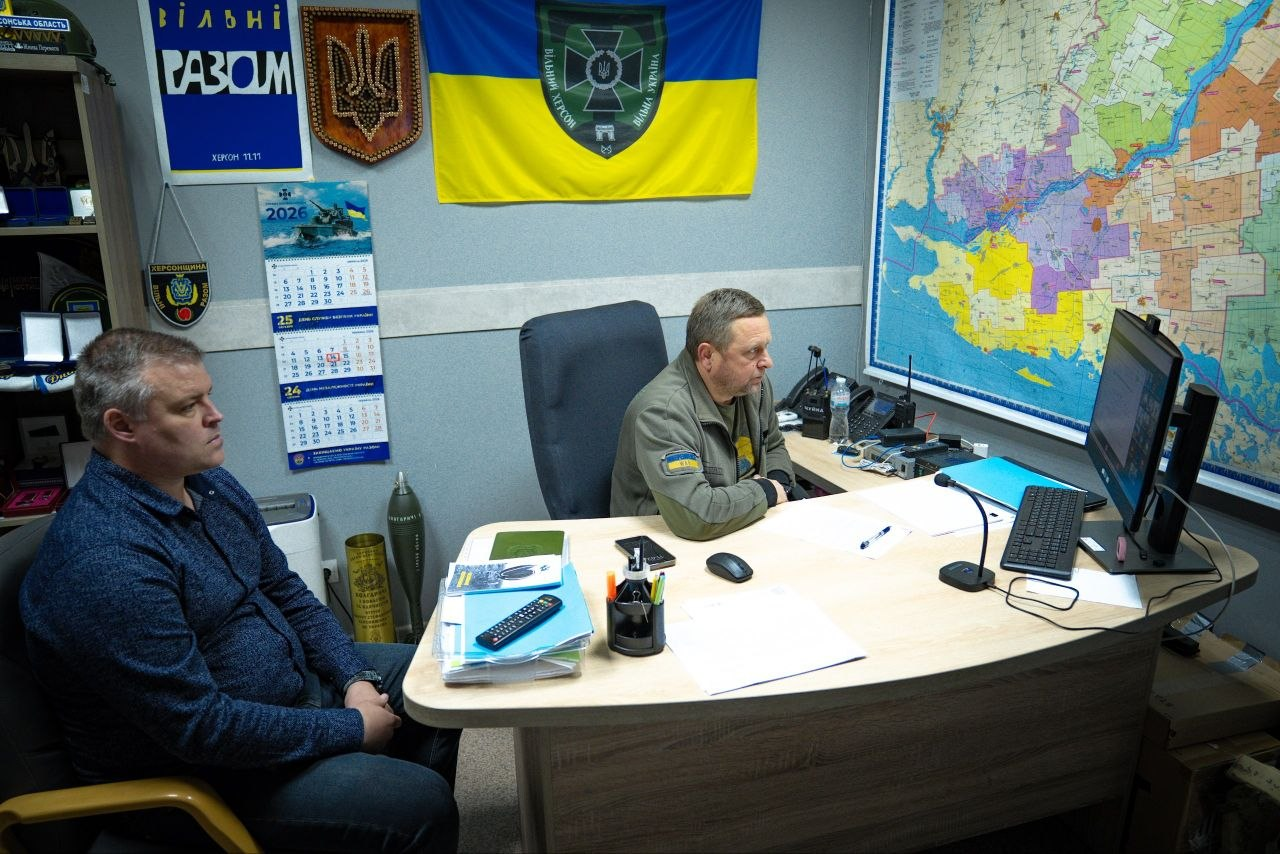 Двоє чоловіків перебувають у робочому кабінеті. Один сидить за столом перед монітором комп'ютера, інший — навпроти нього на стільці. На стінах кімнати розміщено прапор України з емблемою, велику карту, державний герб, патріотичні плакати та настінний календар.