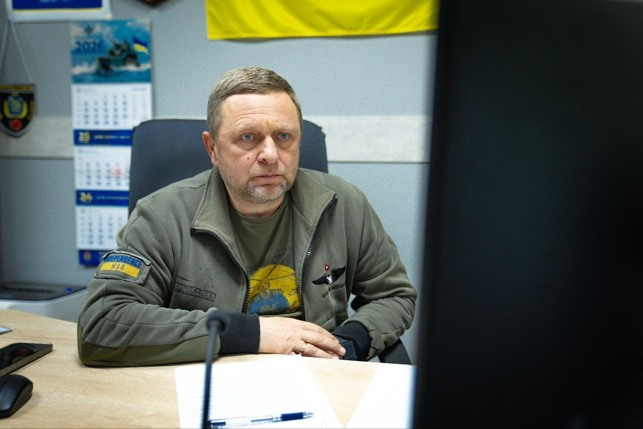 Чоловік у зеленій куртці мілітарі сидить за робочим столом в офісі та дивиться у бік комп'ютерного монітора, який частково видно на передньому плані. На його куртці є нашивки, а на стіні на задньому плані висить настінний календар та частина прапора України.