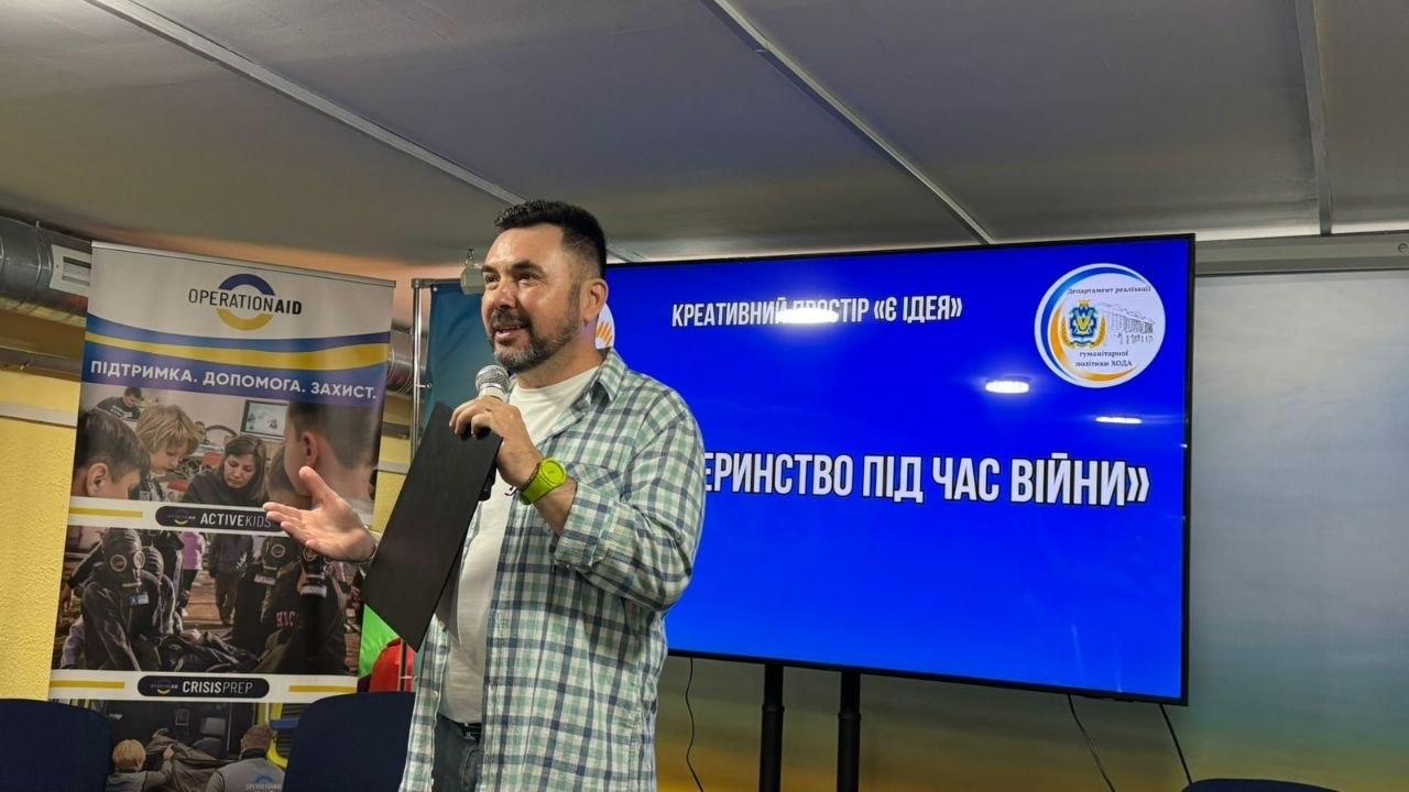 Чоловік у кардигані в клітинку виступає з мікрофоном та папкою у руках перед аудиторією. За ним розташований великий синій екран із логотипом та текстом «Креативний простір 