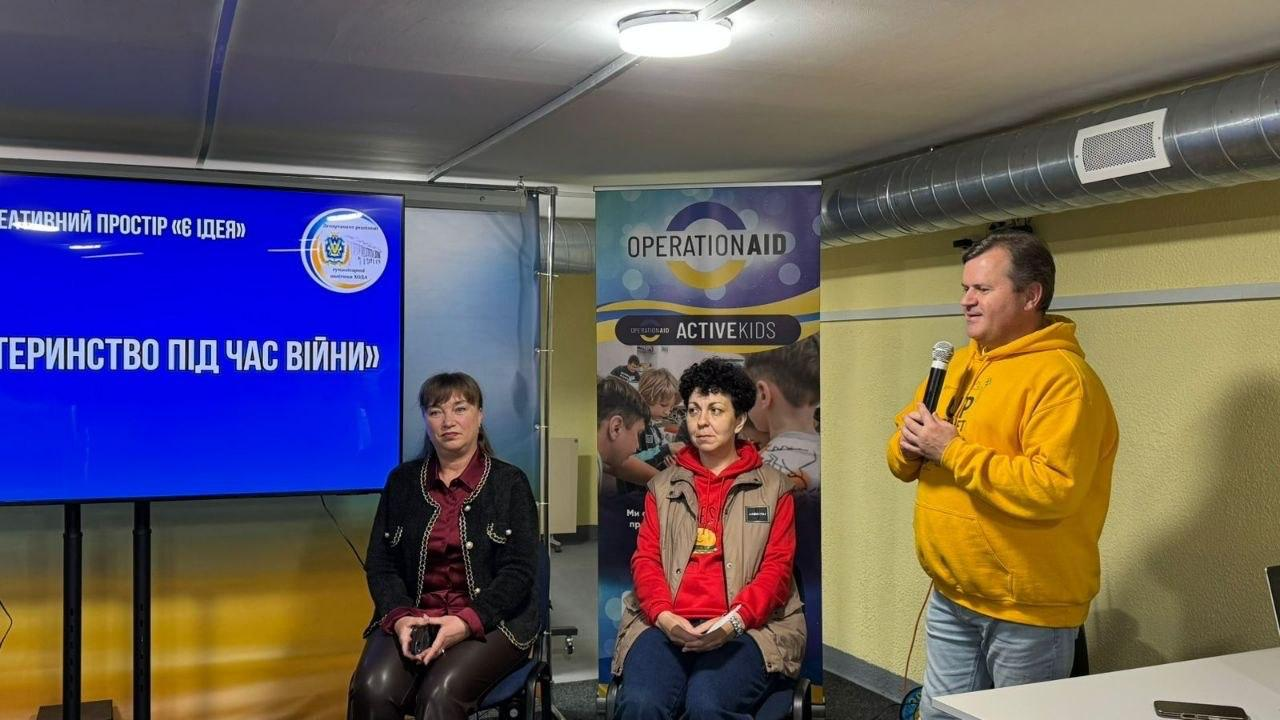 Чоловік у жовтому худі виступає з мікрофоном у руках перед аудиторією. Поруч на стільцях сидять дві жінки та слухають його. На задньому плані розміщено великий синій екран із написом «Материнство під час війни» та вертикальний банер організації «Operation Aid».