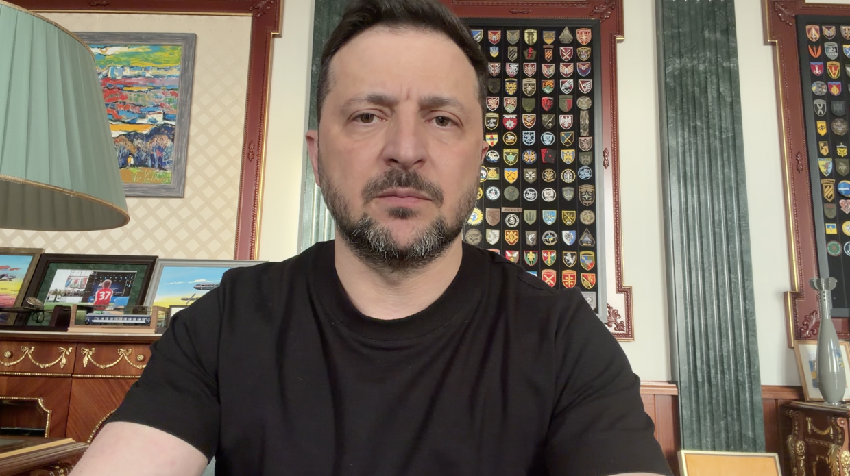 Чоловік у чорній футболці зображений крупним планом на тлі офісного інтер'єру. На стіні позаду нього висить велика колекція військових шевронів у рамці, а також художня картина. Ліворуч на комоді стоять невеликі фотокартки та настільна лампа з зеленим абажуром.