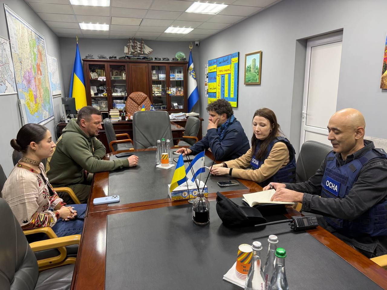 Група людей сидить за великим столом у кабінеті під час офіційної зустрічі. З одного боку столу сидять двоє представників приймаючої сторони, з іншого — троє представників міжнародної місії у захисних жилетах з написом «OCHA». На столі стоять маленькі прапорці, а на стінах висять карти та прапори.