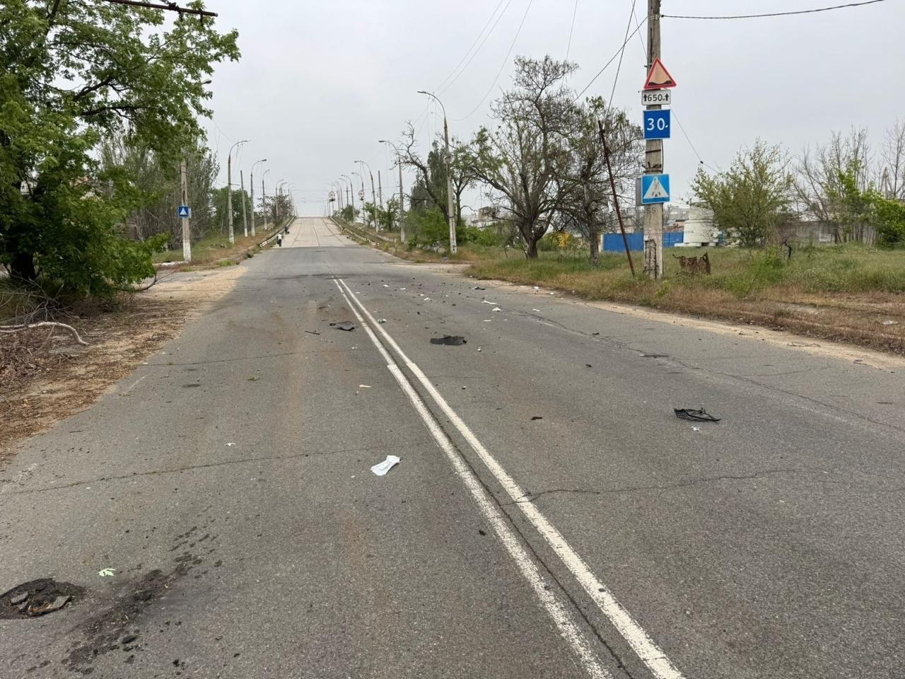 Дволінійна асфальтована дорога з вибоїнами та дрібними уламками на поверхні веде вгору до мосту. Уздовж узбіччя розташовані зелені дерева, стовпи електропередач та дорожні знаки обмеження ваги й швидкості.