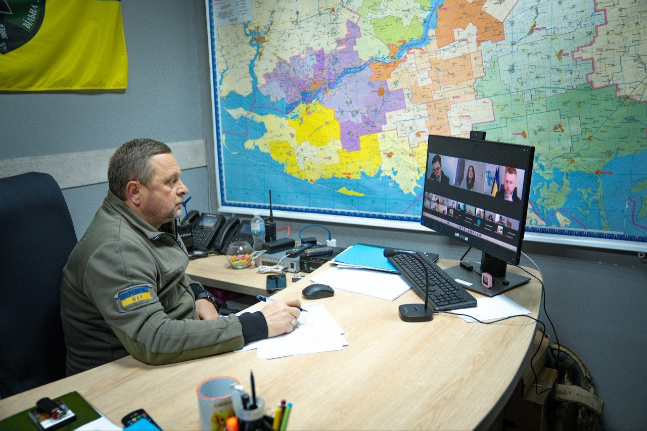 Чоловік у військовій формі сидить за робочим столом перед монітором комп'ютера під час онлайн-наради, на стіні позаду висить велика карта.