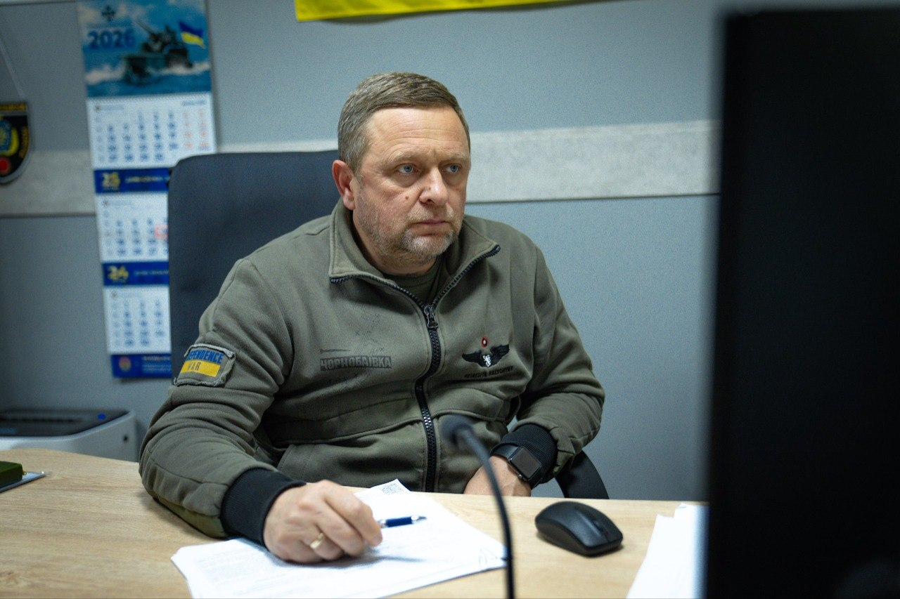 Чоловік у військовій формі сидить за робочим столом перед монітором комп'ютера, тримаючи ручку біля документів.