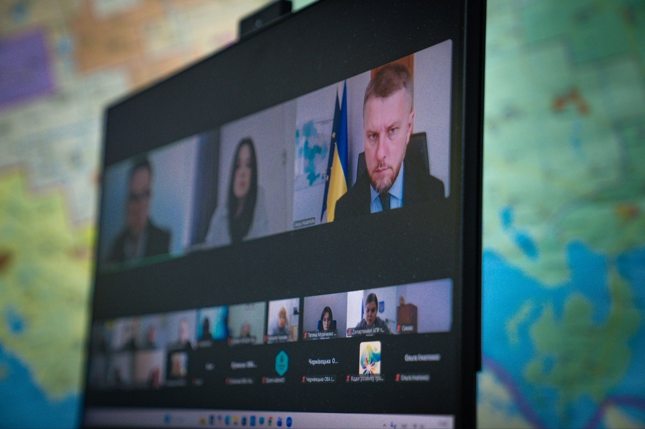 Екран комп'ютерного монітора з вікнами учасників онлайн-наради на тлі розмитої карти на стіні.