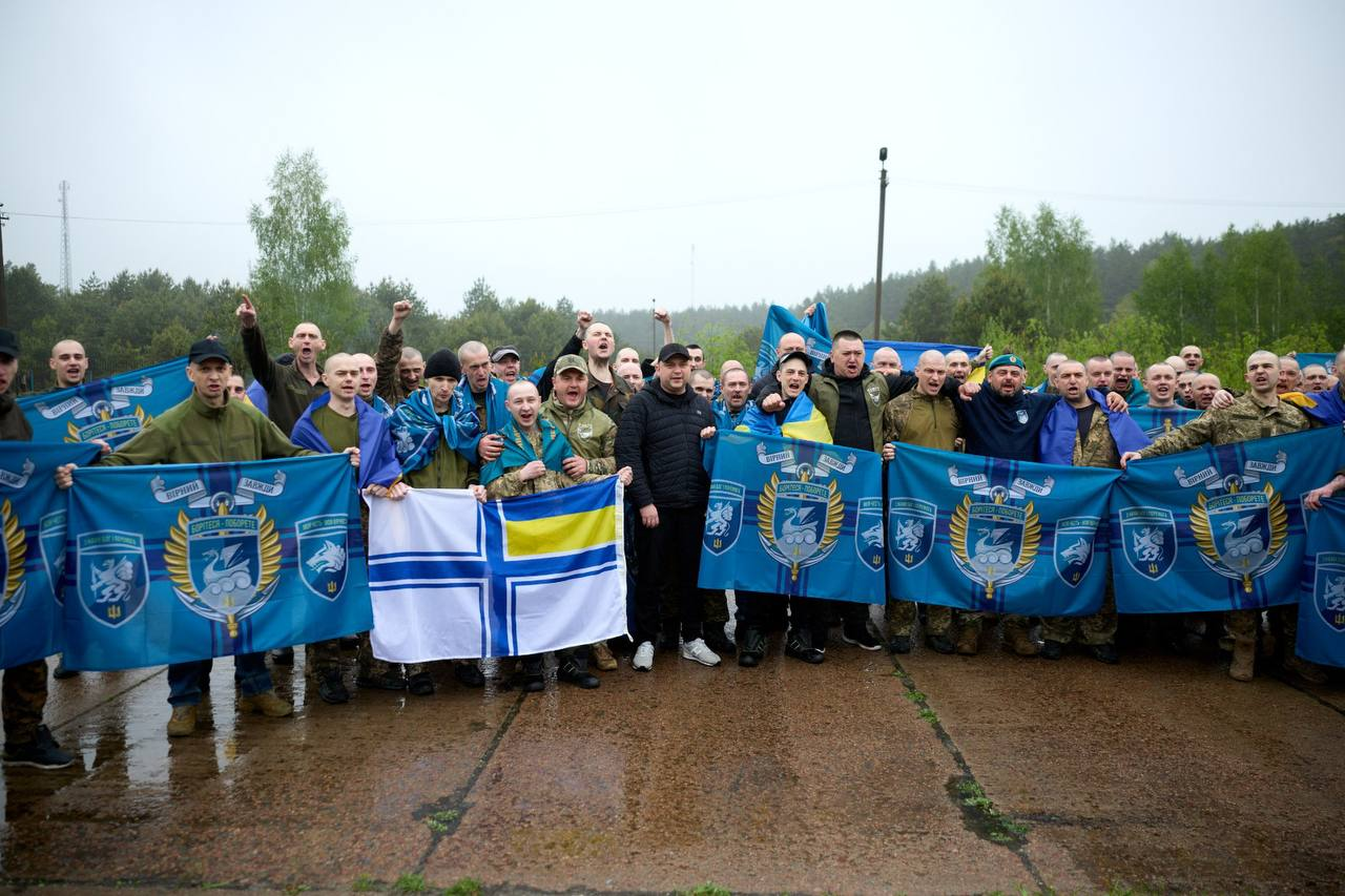 Велика група чоловіків, частина з яких у військовій формі, радісно позує на відкритому повітрі, тримаючи прапори та військову символіку на тлі лісу.