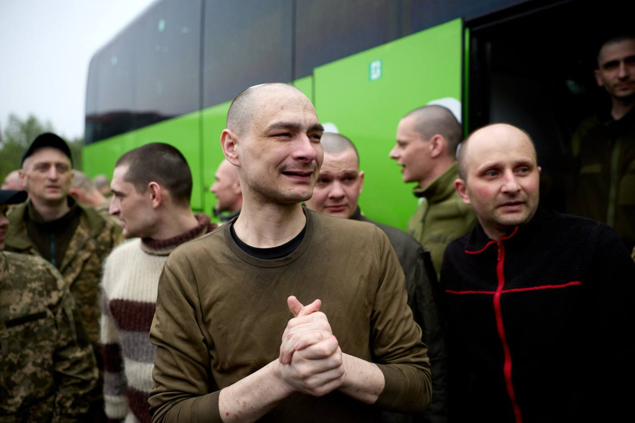 Група чоловіків із поголеними головами, серед яких військові у камуфляжі, стоїть на вулиці біля великого зеленого автобуса. Один із чоловіків на передньому плані емоційно стискає руки разом із виразом радості на обличчі.