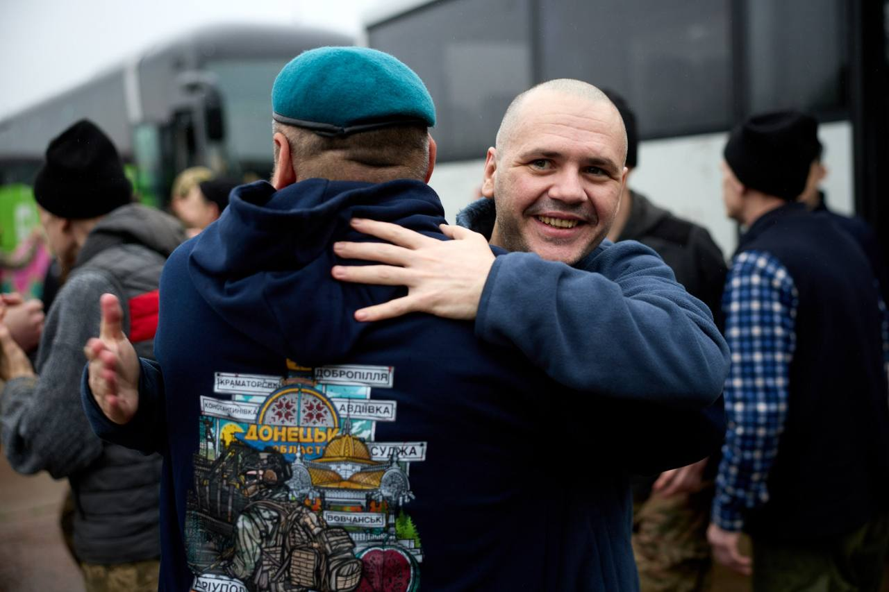Чоловік із поголеною головою радісно посміхається, обіймаючи іншого чоловіка в синьому худі з патріотичним принтом та береті на тлі автобуса.