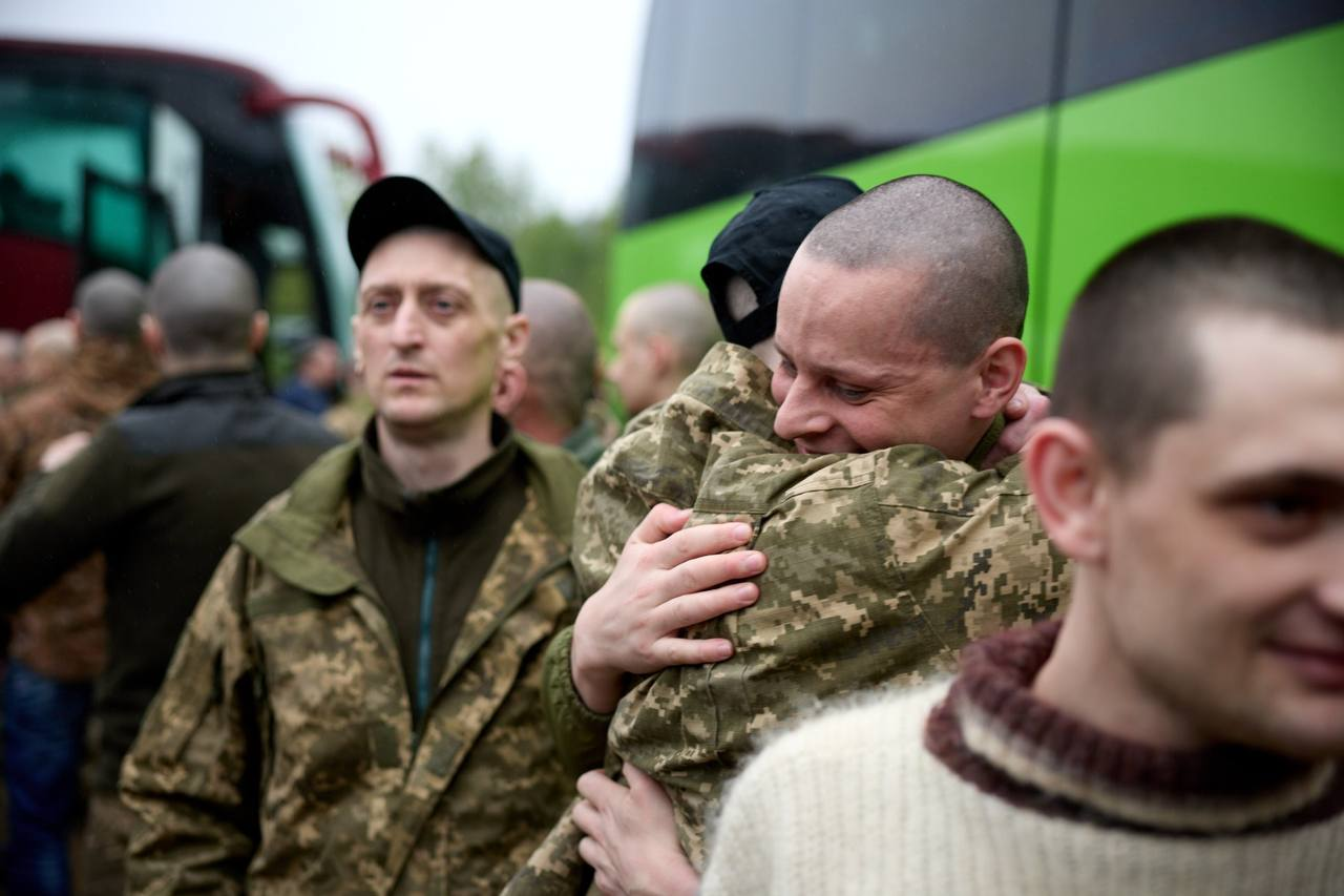 Чоловік у військовій формі емоційно обіймає іншу людину на тлі великого зеленого автобуса, поруч стоять інші чоловіки у цивільному одязі та камуфляжі.