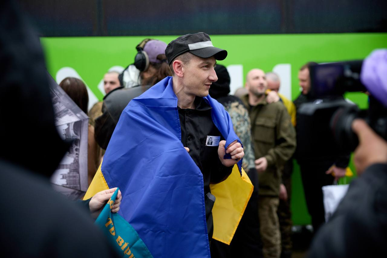 Чоловік у чорній кепці та куртці з бейджем загорнутий у синьо-жовтий прапор України і стоїть серед людей на тлі яскраво-зеленого автобуса.