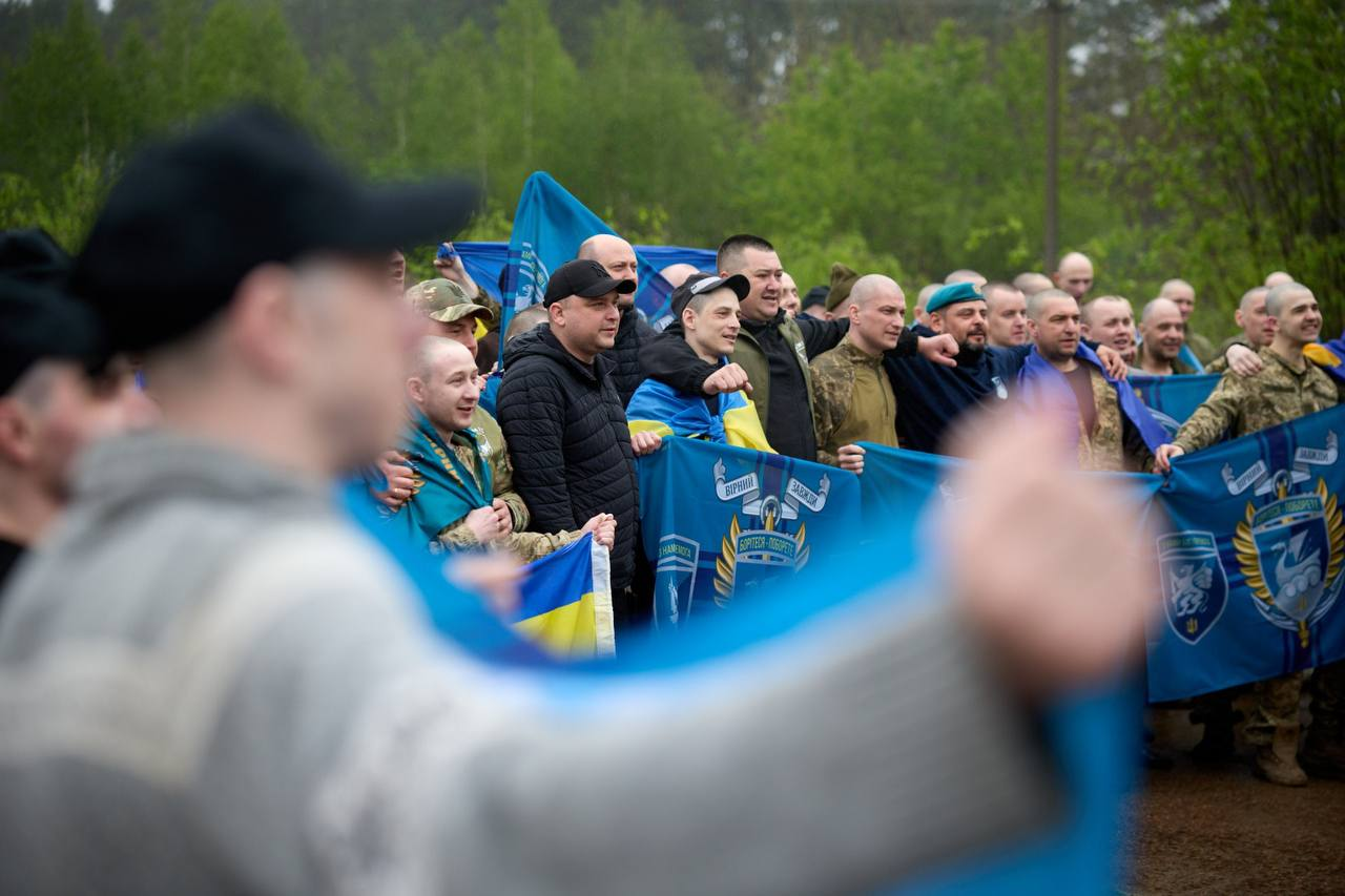 Група чоловіків, серед яких люди у військовій формі, радісно позує на тлі лісу, тримаючи великі сині прапори з емблемами та національну символіку України.