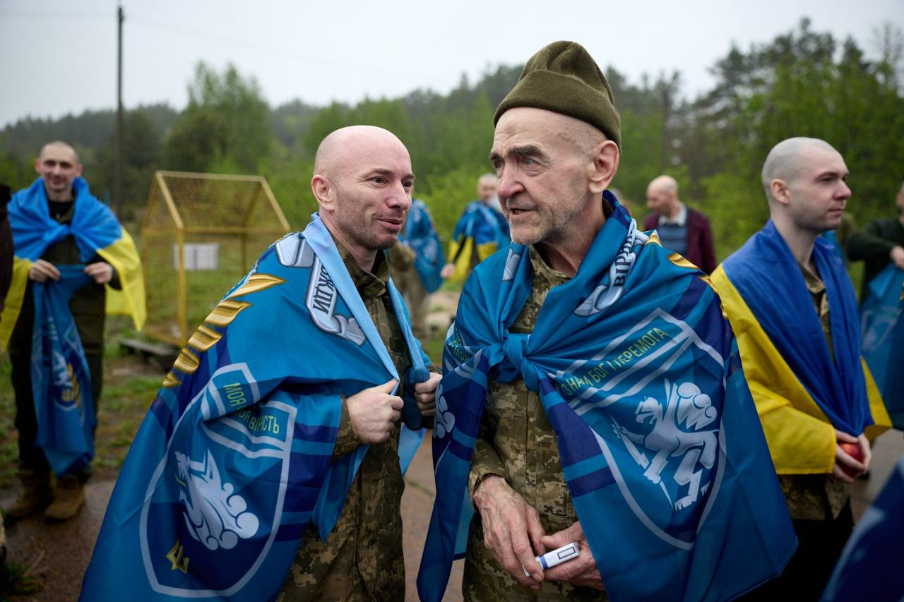 Два чоловіки у військовій формі — один молодшого, а інший старшого віку — спілкуються на вулиці, накинувши на плечі сині прапори з військовою символікою на тлі лісу та інших людей.