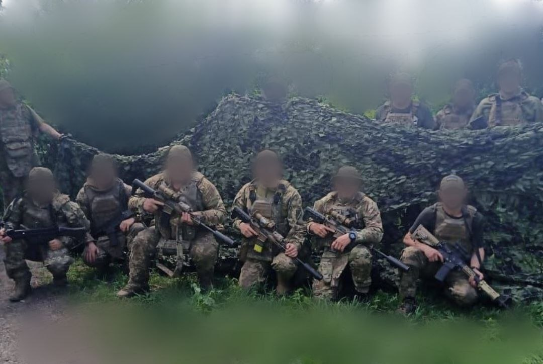 Група військовослужбовців у повному екіпіруванні та зі зброєю позує на тлі великої захисної маскувальної сітки серед зеленої рослинності.