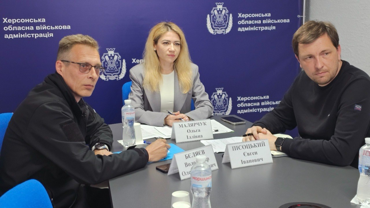 Жінка та двоє чоловіків у цивільному одязі сидять за круглим столом під час офіційного засідання на тлі синього брендованого банера військової адміністрації. Перед кожним із учасників стоять настільні таблички з іменами, папери для записів та пляшки з водою.