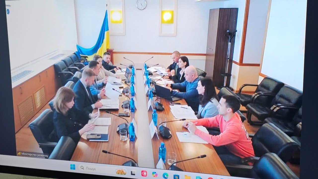 Група людей у цивільному одязі сидить за довгим Т-подібним столом під час засідання у конференц-залі. На столі розкладено папери, ноутбуки, мікрофони та пляшки з водою, а в кутку кімнати стоїть державний прапор України.