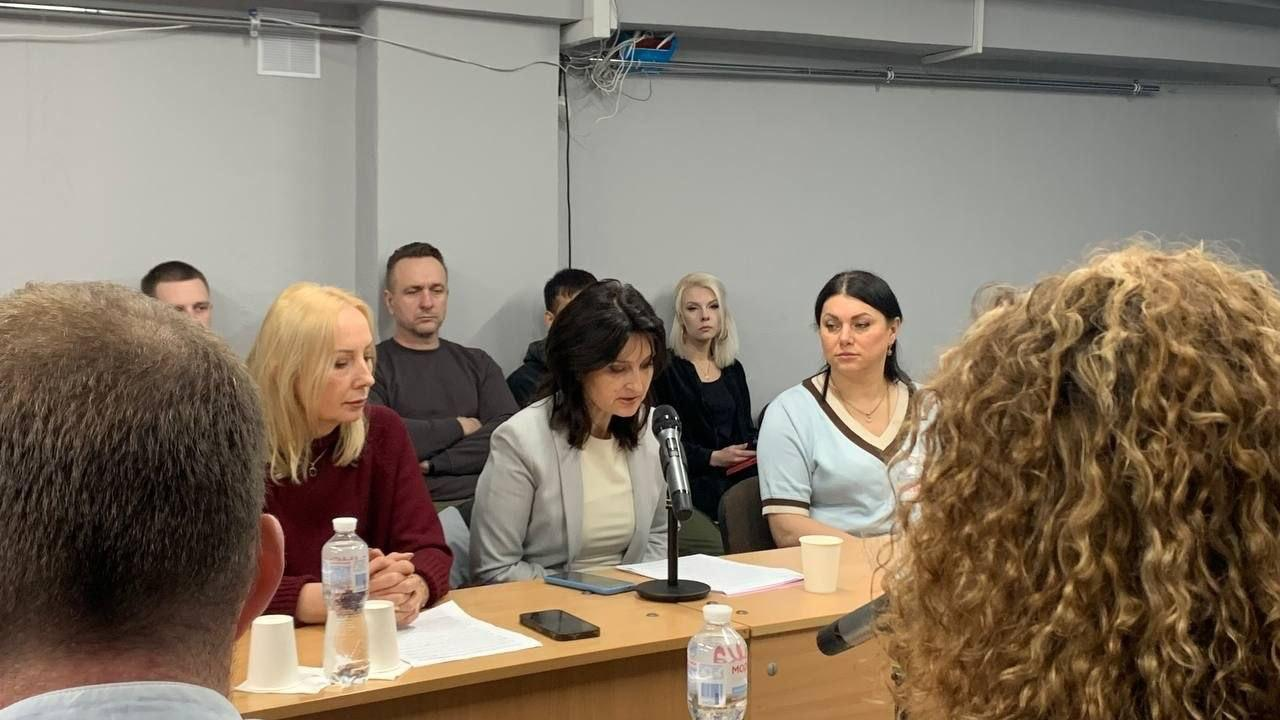 Жінка у світлому піджаку виступає перед мікрофоном під час засідання круглого столу, інші учасники за столом та на задньому плані уважно слухають її.