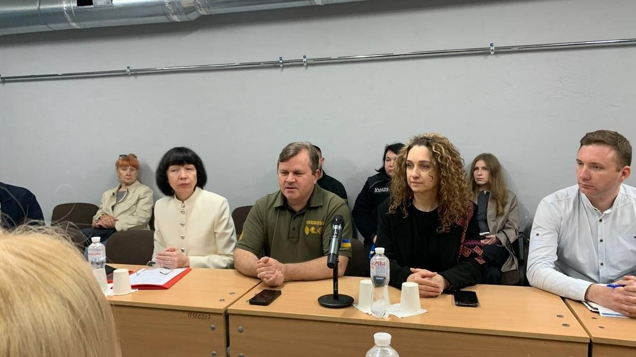 Учасники засідання круглого столу сидять за довгим столом у приміщенні з сірими стінами та трубою на задньому плані. Один із чоловіків у центрі, одягнений у зелену футболку-поло, виступає перед встановленим мікрофоном, тоді як інші присутні уважно слухають.