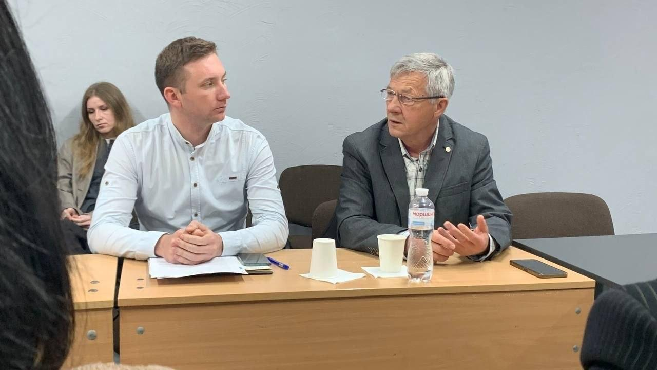 Старший чоловік у піджаку та окулярах активно виступає під час засідання круглого столу, жестикулюючи рукою, тоді як молодий чоловік у світлій сорочці поруч уважно його слухає.