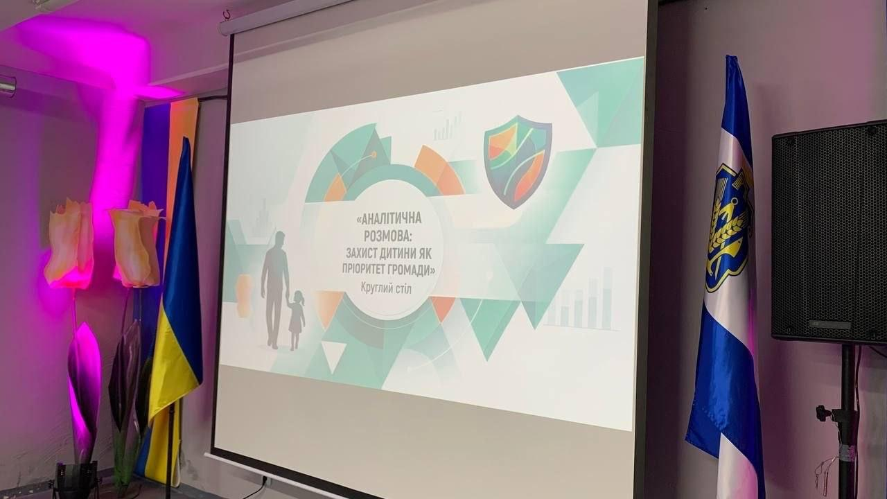 Великий проекційний екран із головним слайдом круглого столу «Аналітична розмова: захист дитини як пріоритет громади» в залі з прапорами та декоративним підсвічуванням.