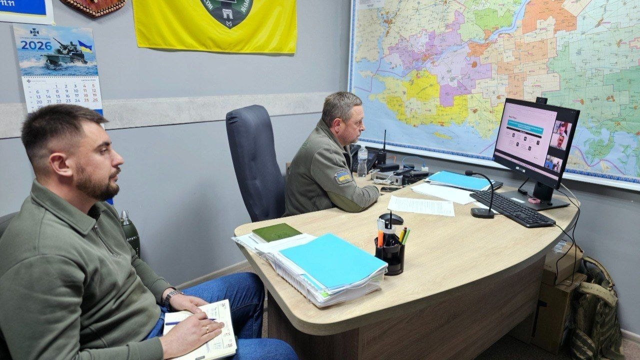 Два чоловіки у військовому та цивільному одязі проводять онлайн-нараду перед монітором комп'ютера у кабінеті з великою картою на стіні.