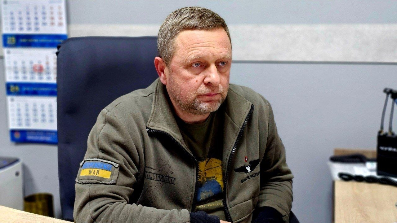Чоловік у військовій кофті з нашивками сидить у кабінеті за робочим столом на тлі настінного календаря.