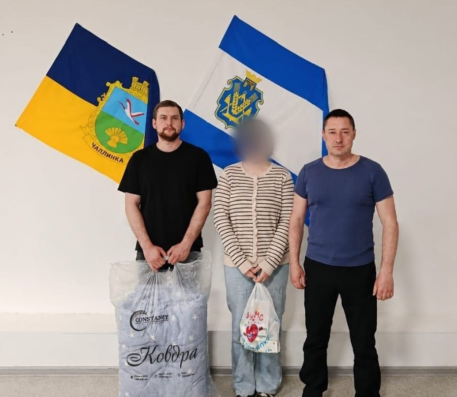 Два чоловіки та жінка із розмитим обличчям стоять у приміщенні на тлі прапорів Чаплинки та Херсонської області. Один із чоловіків тримає великий прозорий пакунок із написом «Ковдра», а жінка тримає в руці білий пакунок із малюнком серця.