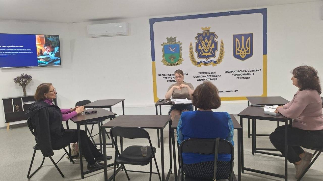 П’ятеро жінок проводять робочу зустріч у кабінеті навколо розставлених окремих столів. На стіні позаду висить великий банер із гербами України, Херсонської області, Чаплинської та Долматівської громад, а ліворуч на стіні закріплено ввімкнений телевізор.