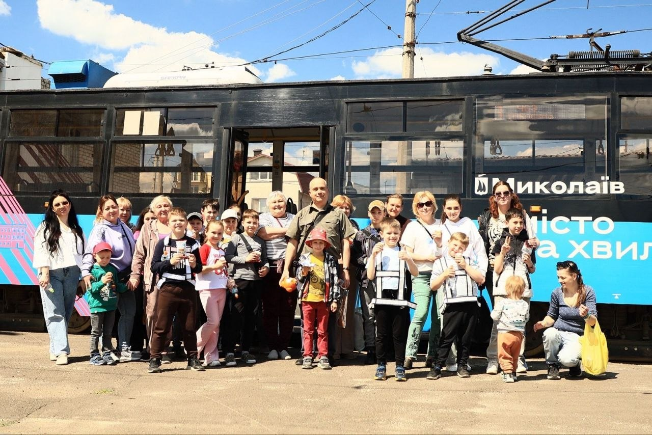 Велика група дорослих та дітей позує для спільного фото на вулиці на тлі синьо-чорного брендованого трамвая з написом «Миколаїв».