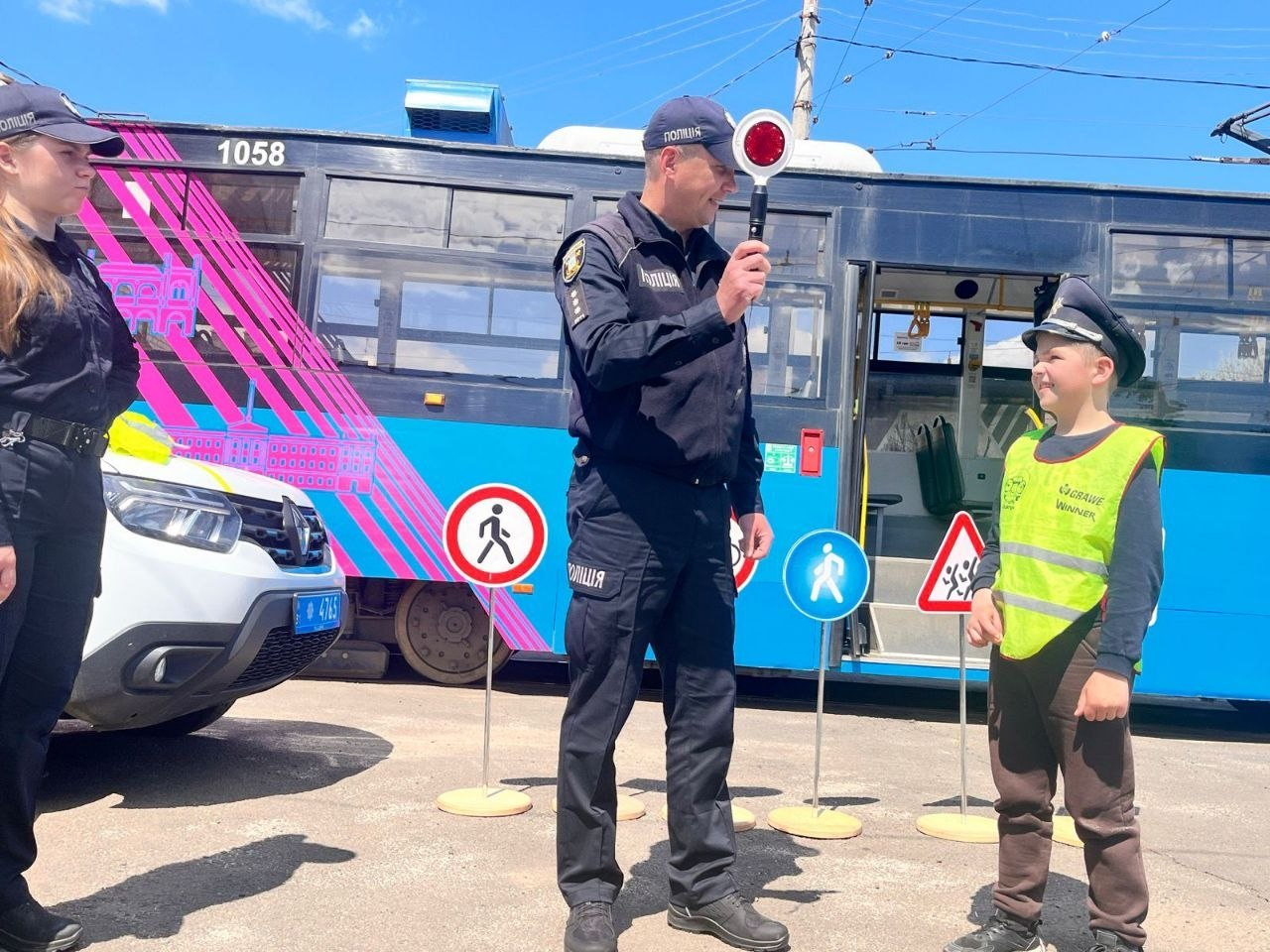 Співробітники поліції проводять навчальне заняття з правил дорожнього руху для хлопчика у світловідбивальному жилеті та кашкеті на тлі патрульного автомобіля і трамвая. Поліцейський демонструє дитині регулювальний диск, а поруч на стійках встановлено макети дорожніх знаків.