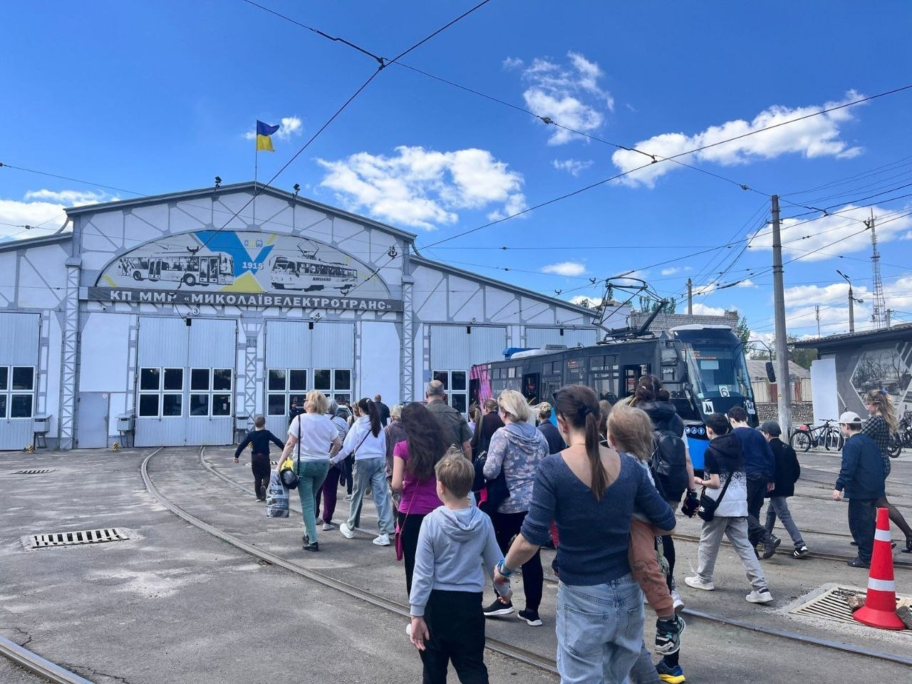 Велика група дітей та дорослих іде територією депо до великої промислової будівлі з відчиненими воротами, поруч на коліях стоїть сучасний трамвай.