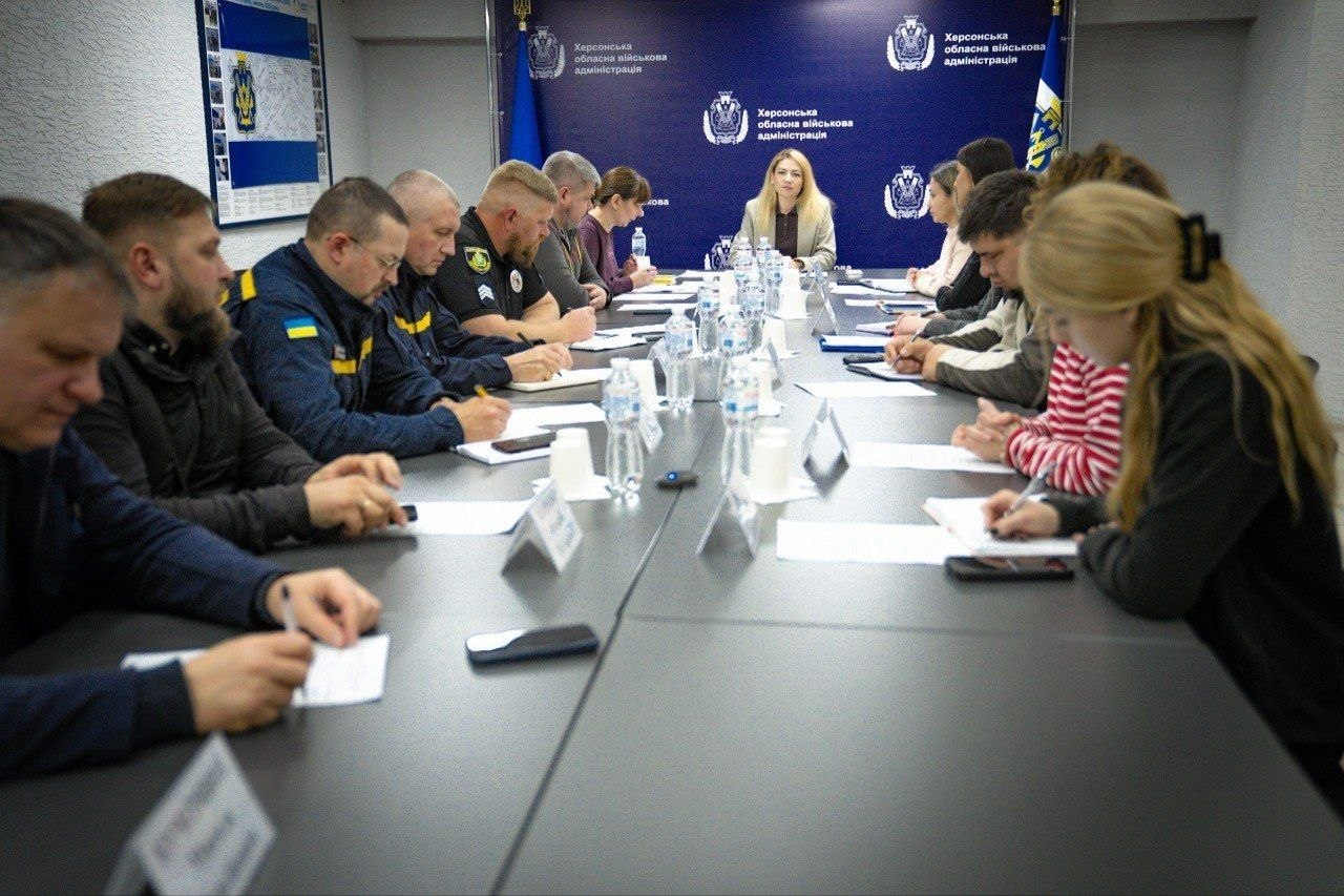 Нарада у конференц-залі, де учасники сидять за довгим столом, роблять нотатки та слухають керівника, який перебуває в торці столу на тлі синього брендованого банера з логотипами.