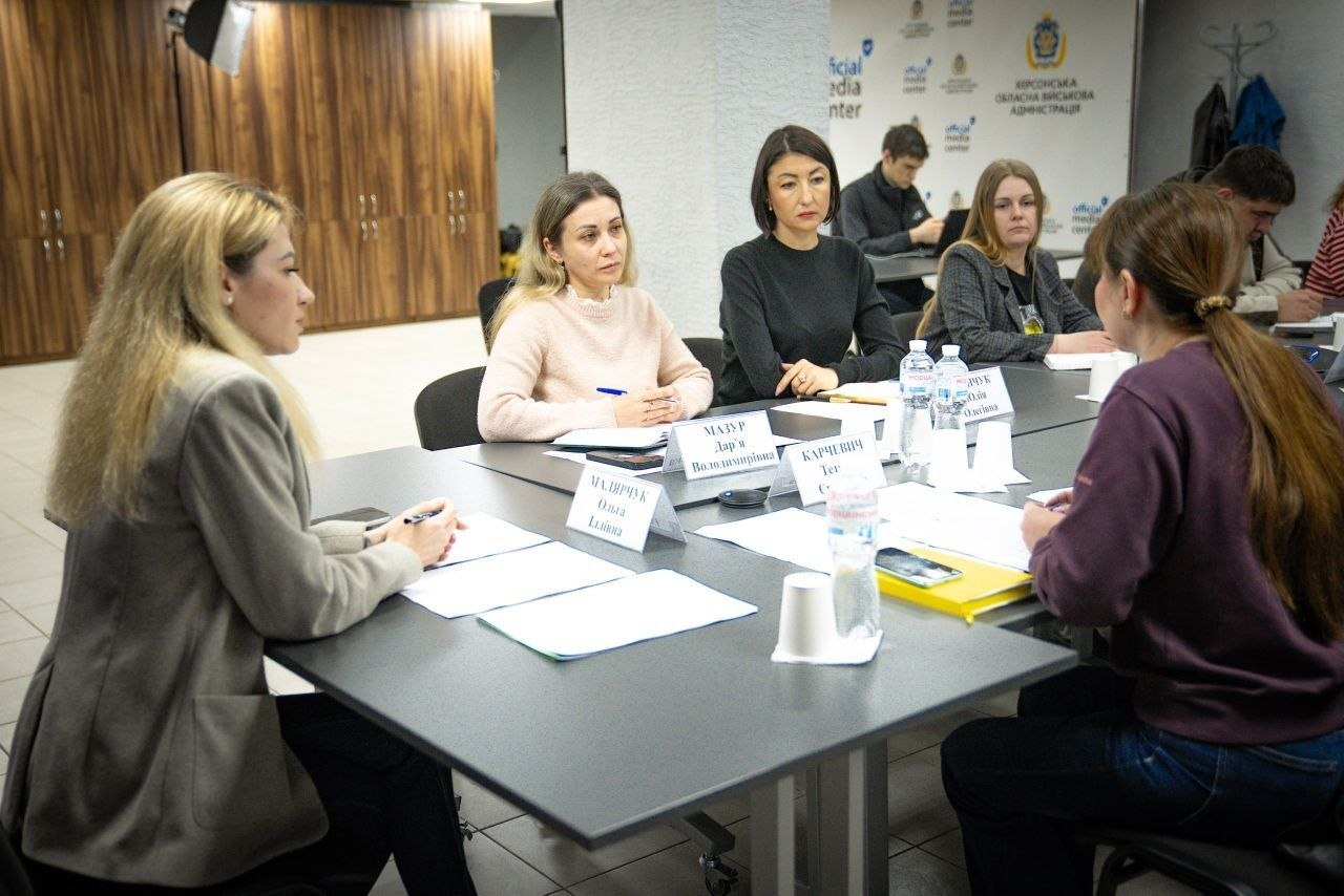 Нарада у конференц-залі за участю керівниці та інших учасниць, які сидять за Т-подібним столом із документами та табличками. Вони спілкуються та обговорюють робочі питання на тлі світлого банера офіційного медіацентру.