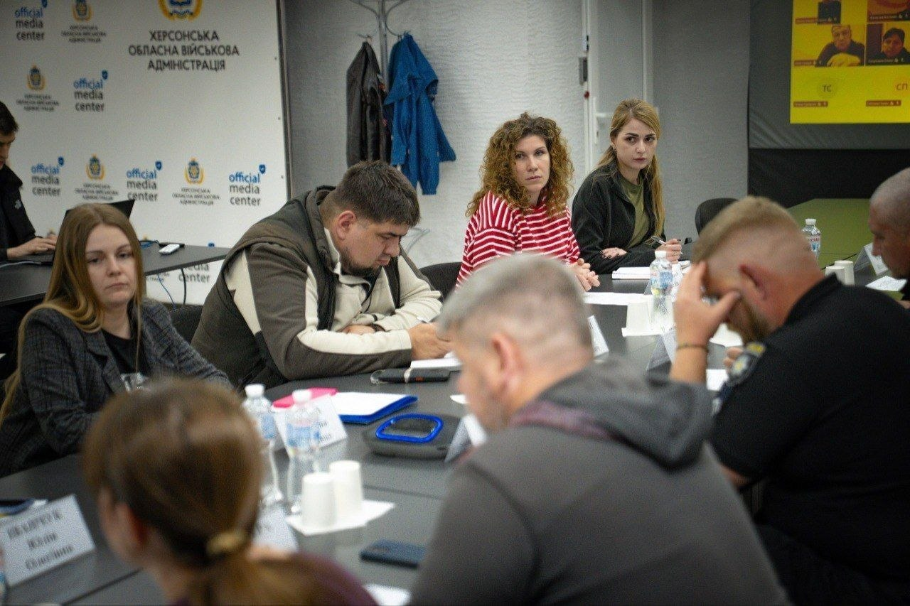 Нарада у конференц-залі за участю представників відомств та служб, які сидять за столом, роблять нотатки та слухають спікера. На задньому плані розміщено світлий брендований банер офіційного медіацентру.
