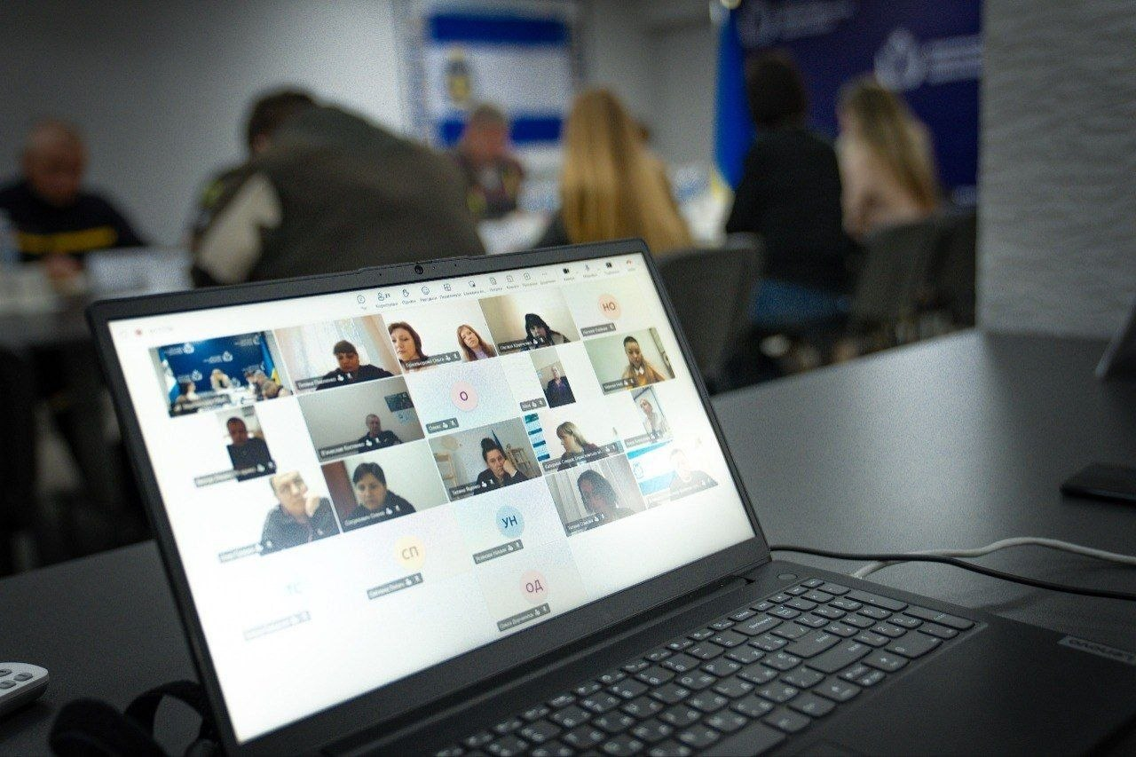 Ноутбук на передньому плані з відкритим вікном онлайн-наради, де на екрані видно сітку з багатьма учасниками відеоконференції. На розмитому задньому плані видно частину конференц-залу, де люди також сидять за столом.