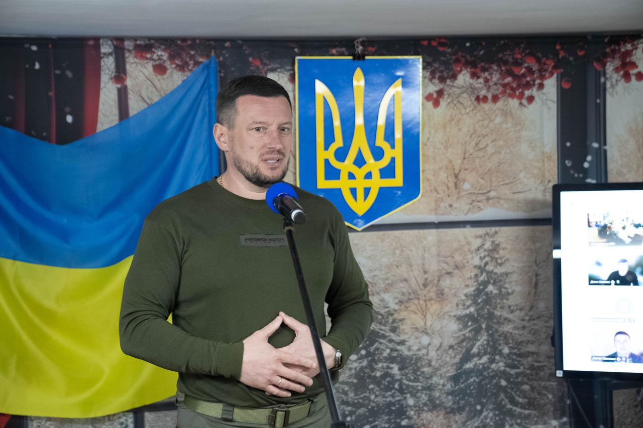 Чоловік у військовому одязі хакі виступає з привітанням перед мікрофоном. Позаду нього розміщено Державний Герб України та синьо-жовтий прапор, а праворуч видно увімкнений екран.