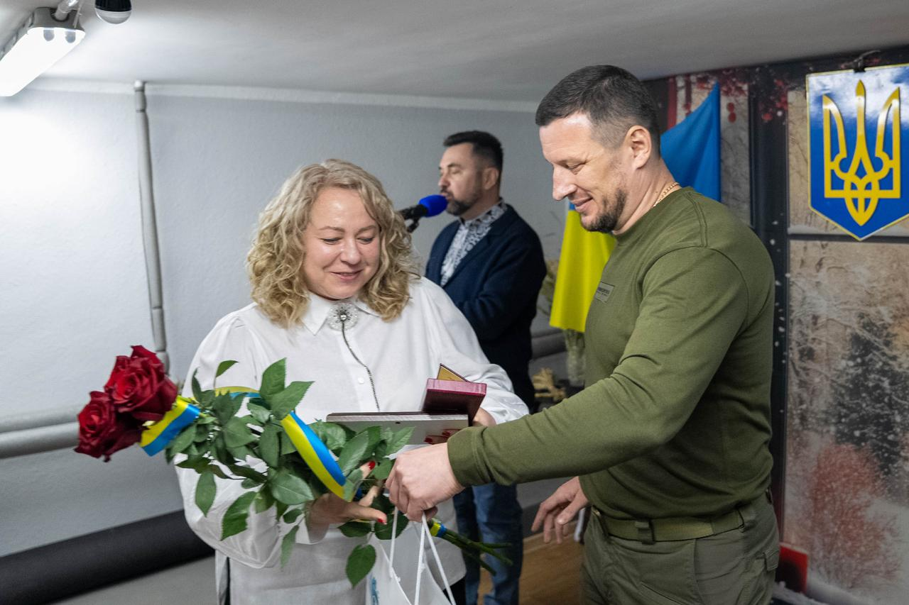 Урочисте вручення нагороди, де чоловік у військовому одязі хакі передає державну відзнаку, білий пакет та букет червоних троянд жінці у білій сорочці. На задньому плані видно ведучого біля мікрофона, а також Державний Герб та синьо-жовтий прапор.